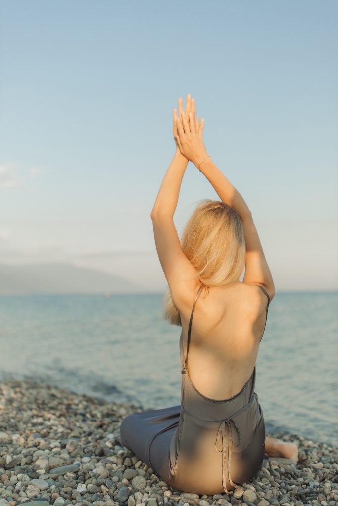 woman does a stretch on a pebble beach. she wears a dress, which exposes her back.