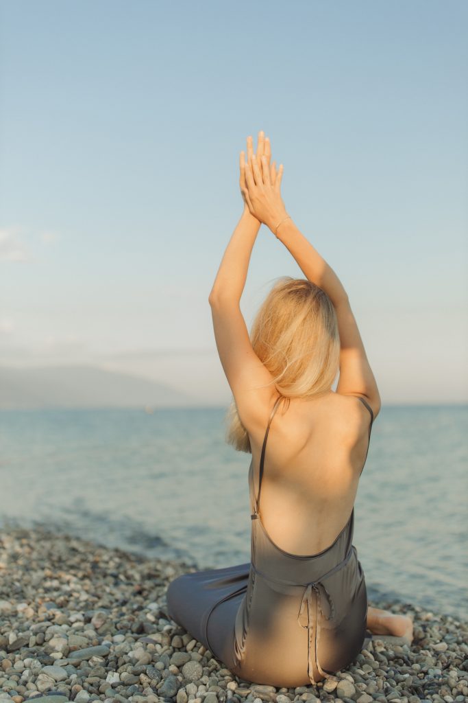 woman does a stretch on a pebble beach. she wears a dress, which exposes her back.