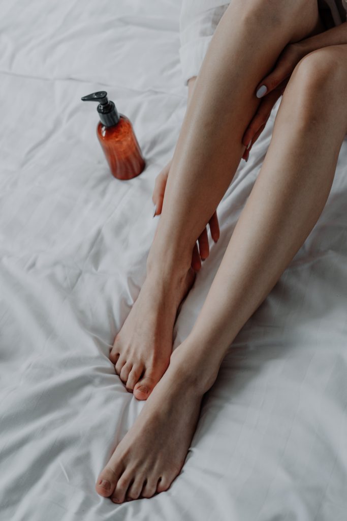 a woman touches her bare legs while laying on a white sheet. there is a pump bottle next to her.